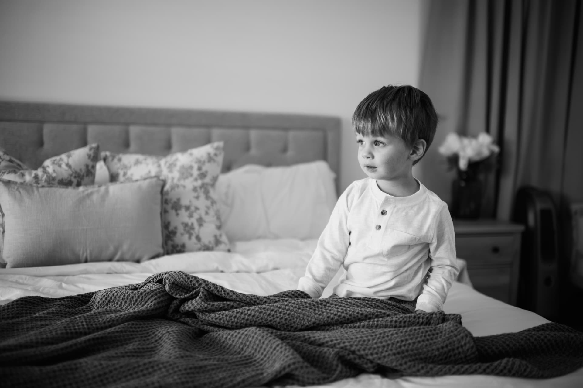 Black and white photo of a little boy sitting on a bed under a knit blanket, gazing toward the window